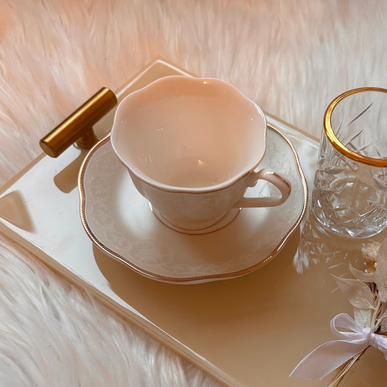 May include: A white porcelain teacup and saucer with a gold trim, sitting on a beige tray with a gold handle. The tray is on a white fluffy surface.