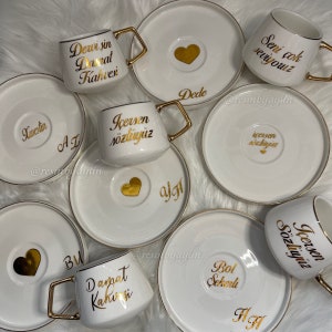 May include: Set of ten white ceramic coffee cups with gold trim and saucers. Each cup has a different gold inscription, including "Dewi sin Danat Kahvesi", "Icersen sozluyuz", "Seni cok seviyouz", "Dede", "Kwestin", "AL", "BU", "algt", "Bol Sekerli", and "HA".