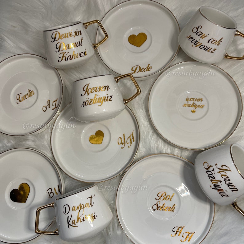May include: Set of ten white ceramic coffee cups with gold trim and saucers. Each cup has a different gold inscription, including "Dewi sin Danat Kahvesi", "Icersen sozluyuz", "Seni cok seviyouz", "Dede", "Kwestin", "AL", "BU", "algt", "Bol Sekerli", and "HA".