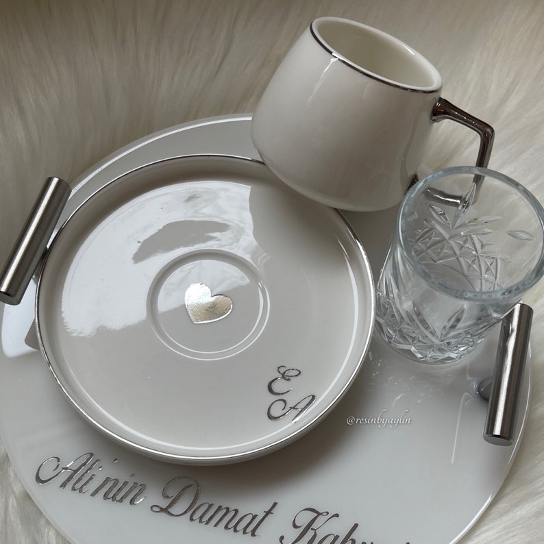 May include: White ceramic tray with silver trim and handles. The tray has a white ceramic cup and saucer with a silver heart and the initials "E & A" on the saucer. There is also a clear glass with a cut design on the tray. The tray has the text "Ali'nin Damat Kah" written on it.