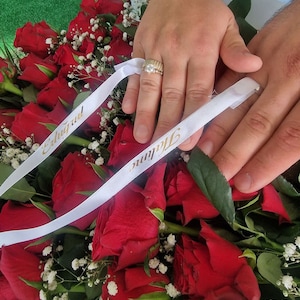 May include: Two hands with wedding rings on their ring fingers, resting on a bouquet of red roses with white baby's breath. The bouquet is tied with two white ribbons with the words "Eduarda" and "Thiago" written in gold.