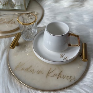 May include: A white marble tray with gold accents and the text "Kadir'in Kahvesi" in gold lettering. The tray has two gold handles and a white ceramic coffee cup and saucer with a gold rim and the initials "G.K." in gold lettering.
