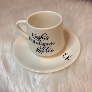 May include: White coffee cup and saucer with black lettering that reads "Kashi's Br&auml;utigam Kaffee" and "EK".