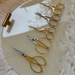 May include: Five gold-colored decorative scissors with ornate handles on a white surface. The scissors are arranged in a vertical line, with the largest scissors at the top and the smallest scissors at the bottom.