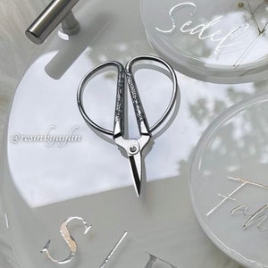May include: A pair of silver-toned, decorative scissors with a heart-shaped design. The scissors are on a white surface with the words "Sealed" and "Faith" written in cursive.