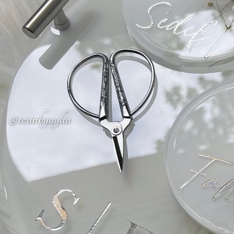 May include: A pair of silver-toned, decorative scissors with a heart-shaped design. The scissors are on a white surface with the words "Sealed" and "Faith" written in cursive.