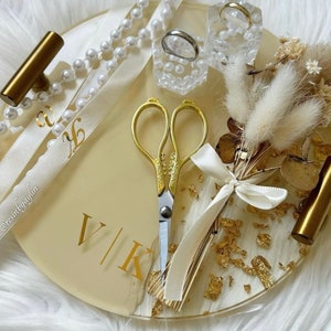 May include: A round, cream-colored tray with gold accents and a gold foil design. The tray has a pair of gold scissors, a small bouquet of dried flowers, two rings in small boxes, and a white ribbon with the text "@restinyagyin" and "V/K".