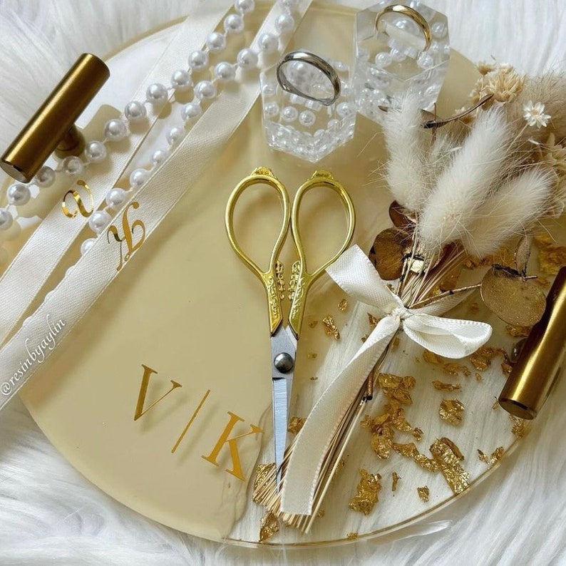 May include: A round, cream-colored tray with gold accents and a gold foil design. The tray has a pair of gold scissors, a small bouquet of dried flowers, two rings in small boxes, and a white ribbon with the text "@restinyagyin" and "V/K".