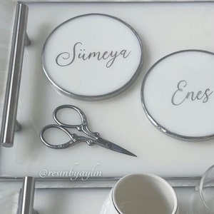 May include: White tray with two round white coasters with silver trim. The coasters have the names "S&uuml;meyra" and "Enes" written in silver script. A pair of silver scissors is on the tray. The tray has a silver handle.