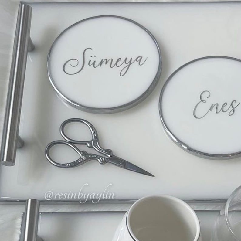 May include: White tray with two round white coasters with silver trim. The coasters have the names "S&uuml;meyra" and "Enes" written in silver script. A pair of silver scissors is on the tray. The tray has a silver handle.