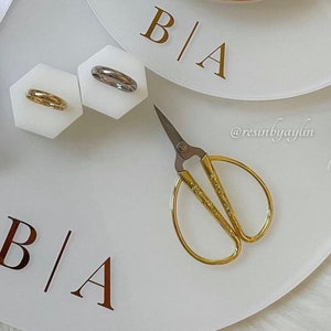 May include: A white tray with the letters "B|A" in gold lettering. The tray has two small white hexagonal dishes with gold and silver rings. A pair of gold scissors is on the tray.