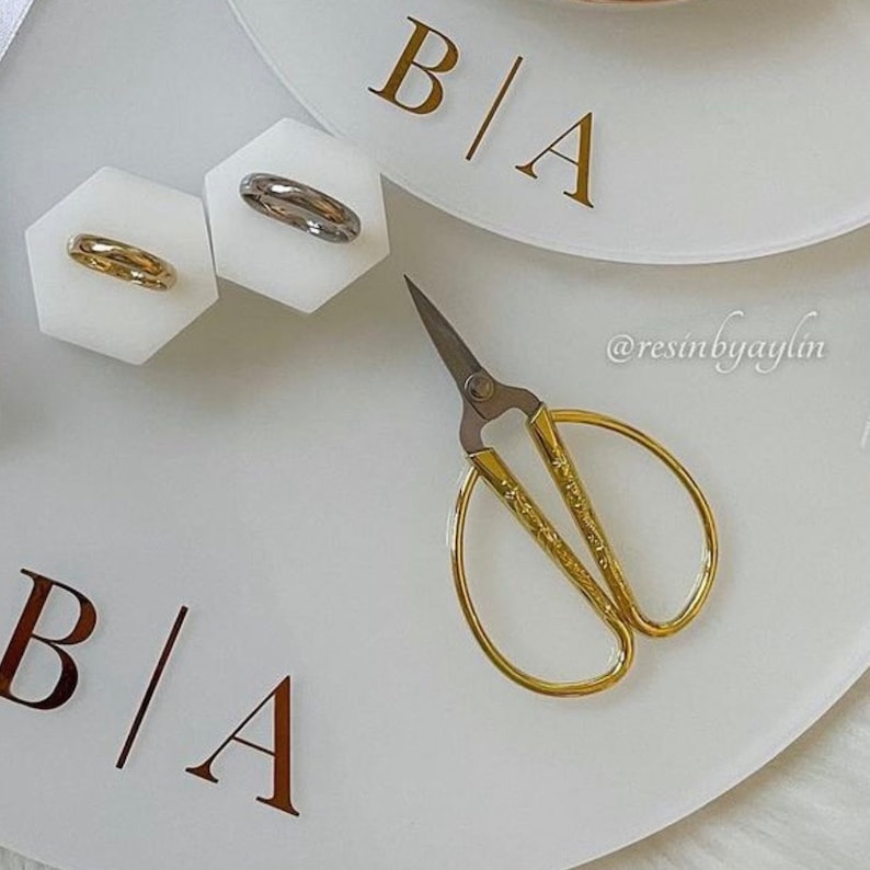May include: A white tray with the letters "B|A" in gold lettering. The tray has two small white hexagonal dishes with gold and silver rings. A pair of gold scissors is on the tray.