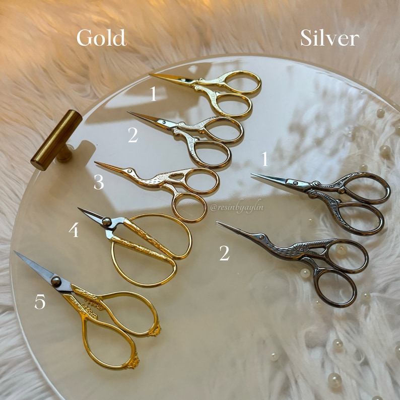 May include: A set of five gold and silver embroidery scissors with bird-shaped handles. The scissors are arranged on a white marble surface with pearl-like accents. The scissors are numbered 1 through 5, with the gold scissors labeled "Gold" and the silver scissors labeled "Silver".