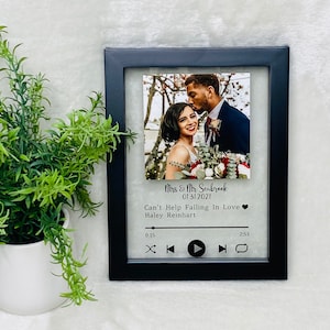 May include: A black framed photo with a wedding picture of a couple in front of a white background. The photo is in a music player frame with the text "Mrs & Mr Seabrook 01 31 2021 Can't Help Falling In Love Haley Reinhart" and a play button.