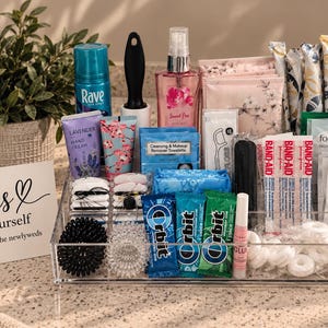 May include: A clear acrylic organizer tray filled with personal care items including Rave hairspray, a lint roller, Sweet Pea body mist, lavender hand cream, makeup remover wipes, dental floss, Band-Aids, Fomin wipes, Shout wipes, Orbit gum, and mints.