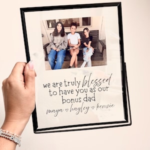 May include: A black rectangular picture frame containing a photograph of three individuals sitting on a porch swing. Below the photo, black text reads: we are truly blessed to have you as our bonus dad, followed by the names maya, hayley, and kenzie.