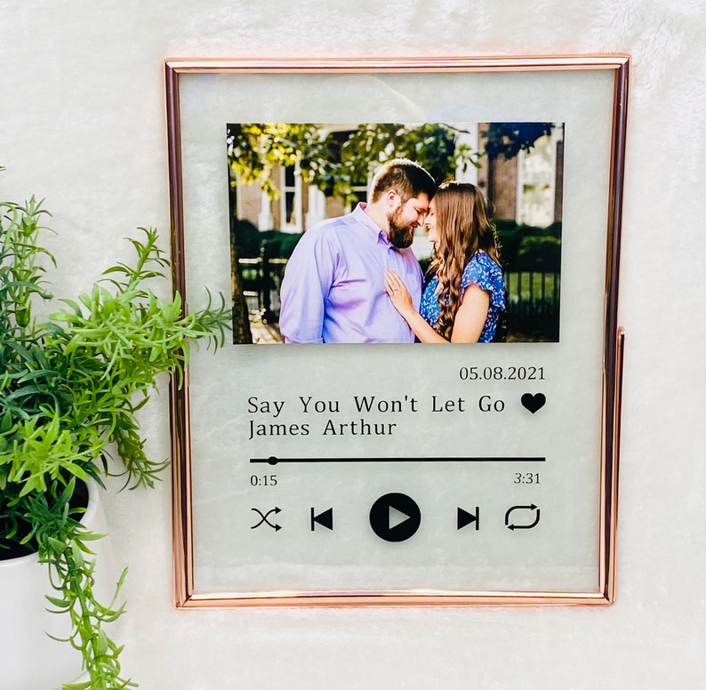 May include: A framed photo with a couple in the background. The photo is in a rose gold frame and has a music player design with the text "Say You Won't Let Go" and "James Arthur" below the photo. The date "05.08.2021" is also included. The music player design includes play, pause, rewind, fast forward, and repeat buttons.
