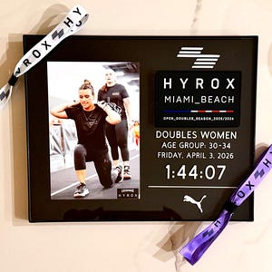 May include: Black framed photo with a picture of two women in athletic wear. The frame includes text: HYROX Miami Beach, Doubles Women, Age Group 30-34, Friday, April 3, 2026, and a time of 1:44:07. Two wristbands with "VROX HY" text.