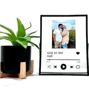 May include: Black framed photo with a couple in a field, a music player graphic with the text "sun to me mgk" and a heart icon. The music player graphic has a play button, rewind button, fast forward button, and a repeat button. The time is 0:15 and 2:38.