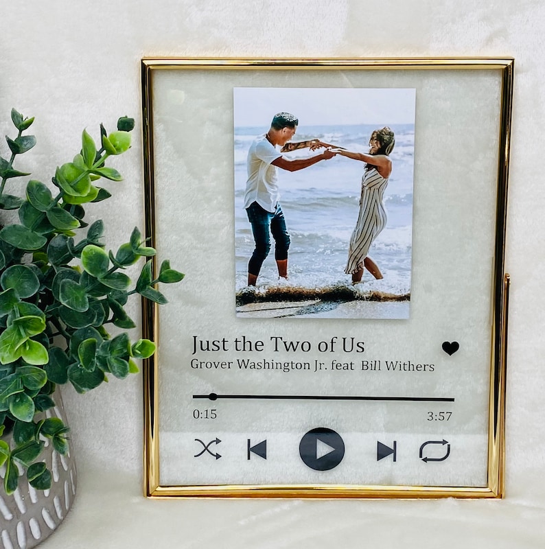 May include: Gold picture frame with a photo of a couple dancing on a beach. The frame has a music player design with the text "Just the Two of Us" and "Grover Washington Jr. feat Bill Withers" and a heart icon.