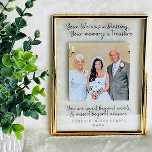 May include: A gold-framed picture frame with a black and white photo of a bride, groom, and another person. The frame has a handwritten message that reads "Your life was a blessing, Your memory a Treasure. You are loved beyond words, & missed beyond measure. FOREVER IN OUR HEARTS PAPA."