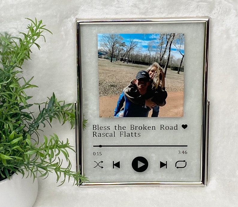 May include: A silver picture frame with a white background and a photo of a couple in a park. The frame has a music player design with the song title "Bless the Broken Road" by Rascal Flatts.