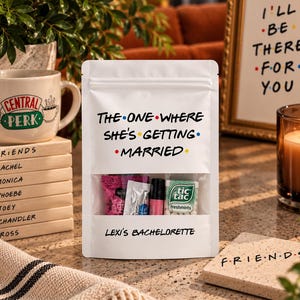 May include: A white stand-up pouch with a clear window containing small items, featuring the text "THE ONE WHERE SHE'S GETTING MARRIED" and "LEXI'S BACHELORETTE" in a black font with colorful dots between the words.