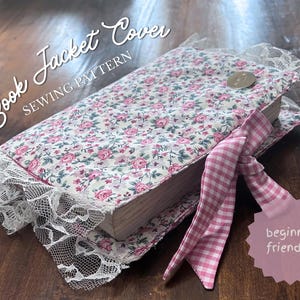 May include: A fabric book jacket cover featuring a floral pattern with pink roses on a cream background, accented by white lace trim on the edges and a pink and white gingham ribbon tie. The text reads Book Jacket Cover SEWING PATTERN and beginner friendly!