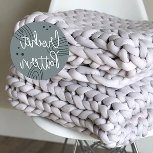 May include: A large, chunky knit throw blanket in a light gray color. The blanket is made with thick yarn and has a woven texture. The blanket is folded and draped over a white chair.