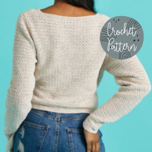 Pu&ograve; includere: Un primo piano di un maglione bianco all'uncinetto con scollo a V e maniche lunghe. Il maglione &egrave; indossato da una persona con jeans blu. Il testo "Crochet Pattern" &egrave; scritto in un cerchio sull'immagine.