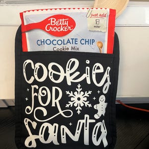 May include: Black oven mitt with white text that says "Cookies for Santa" with a snowflake and gingerbread man design. A Betty Crocker chocolate chip cookie mix box is in the pocket of the oven mitt.