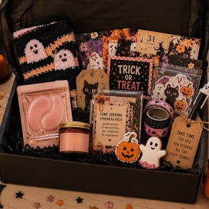 May include: A black gift box filled with Halloween-themed items including fuzzy ghost socks, pink under-eye patches, a small candle, a junk journal ephemera pack, washi tape, a pen, and decorative cutouts featuring pumpkins, ghosts, and a black cat.