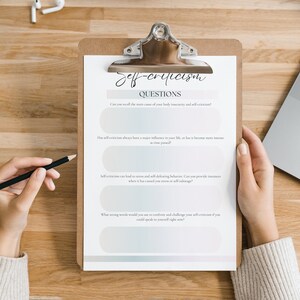 May include: A clipboard with a checklist titled "Self-Criticism Questions". The checklist includes four questions about self-criticism and its impact on life. A person is holding the clipboard and writing on it with a pencil.