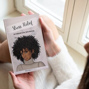 Peut inclure: Une femme tient un journal de travail pour le soulagement du stress avec une illustration color&eacute;e d'une femme sur la couverture. Le texte de la couverture est "Stress Relief Workbook Journal" et "This Girl's Got This".