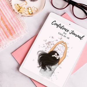 May include: A white tablet with a pink and white striped background. The tablet screen displays a digital journal cover with a girl with long black hair making a heart shape with her hands. The text on the cover reads "Confidence Journal for Girls 10-16".