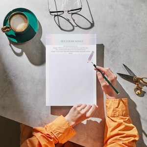 May include: A person in an orange shirt is writing on a white sheet of paper with the words "SET CLEAR GOALS" at the top. The paper is on a gray surface with a green cup of coffee and a pair of glasses.