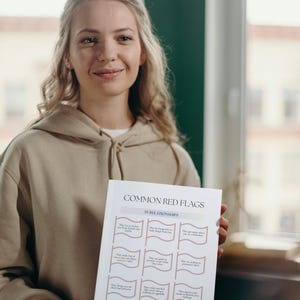 May include: A person holds up a white sheet of paper with the title "Common Red Flags in Relationships". The paper is divided into three columns with nine red flags listed in each column. The red flags are written in black text with a pink flag graphic next to each one.