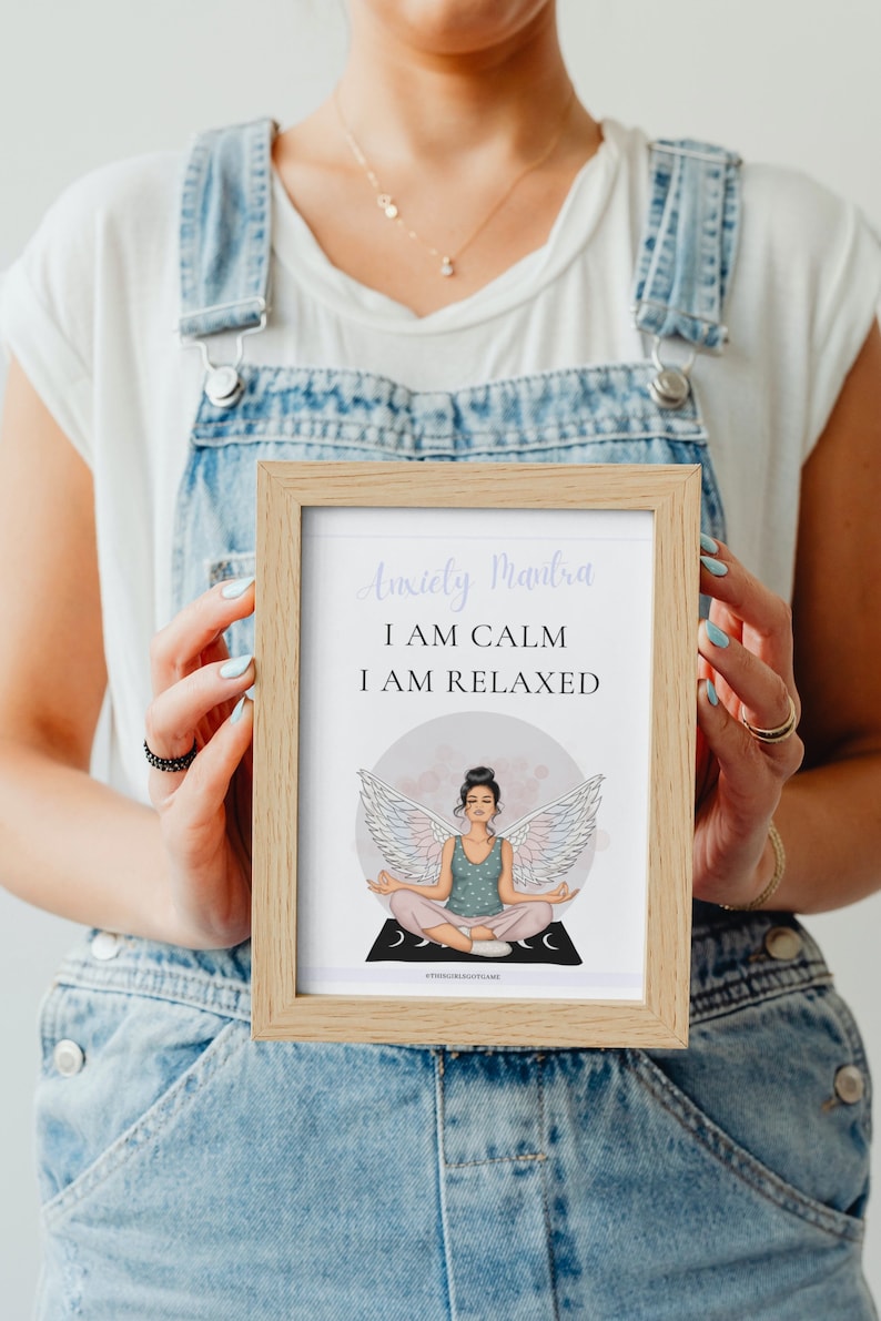 May include: A framed print with a light pink background and a woman with wings meditating. The text reads "Anxiety Mantra I AM CALM I AM RELAXED".