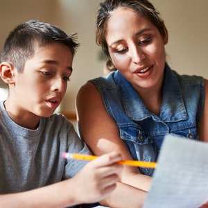 May include: A woman and a boy are sitting at a table, looking at a piece of paper. The boy is holding a pencil and the woman is pointing at the paper. They are likely working on a school assignment together.