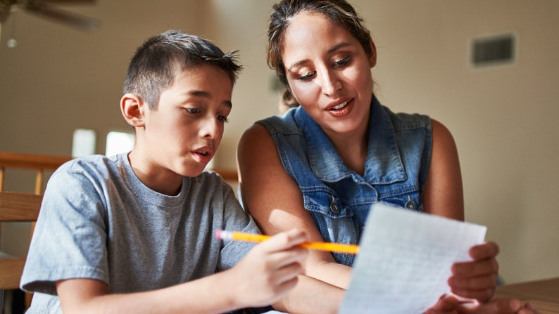 May include: A woman and a boy are sitting at a table, looking at a piece of paper. The boy is holding a pencil and the woman is pointing at the paper. They are likely working on a school assignment together.