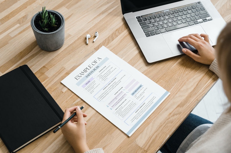 May include: A workspace with a laptop, a printed safety plan, a notebook, and a small potted plant. The safety plan has text and is on a wooden desk. A person is using the laptop and writing on the plan.