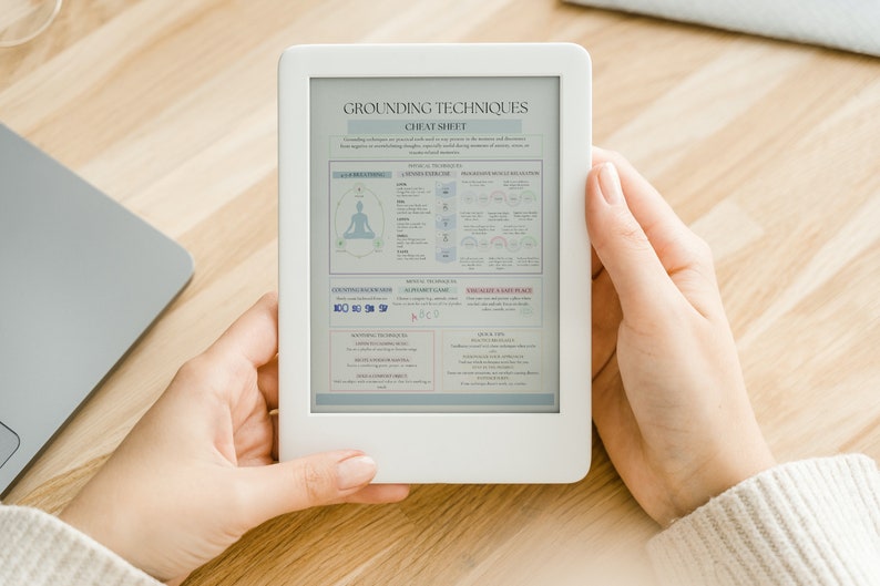 May include: A white e-reader displaying a "Grounding Techniques Cheat Sheet" with text and diagrams. The device is held by a person, with a laptop visible in the background. The e-reader's screen shows information about grounding exercises.