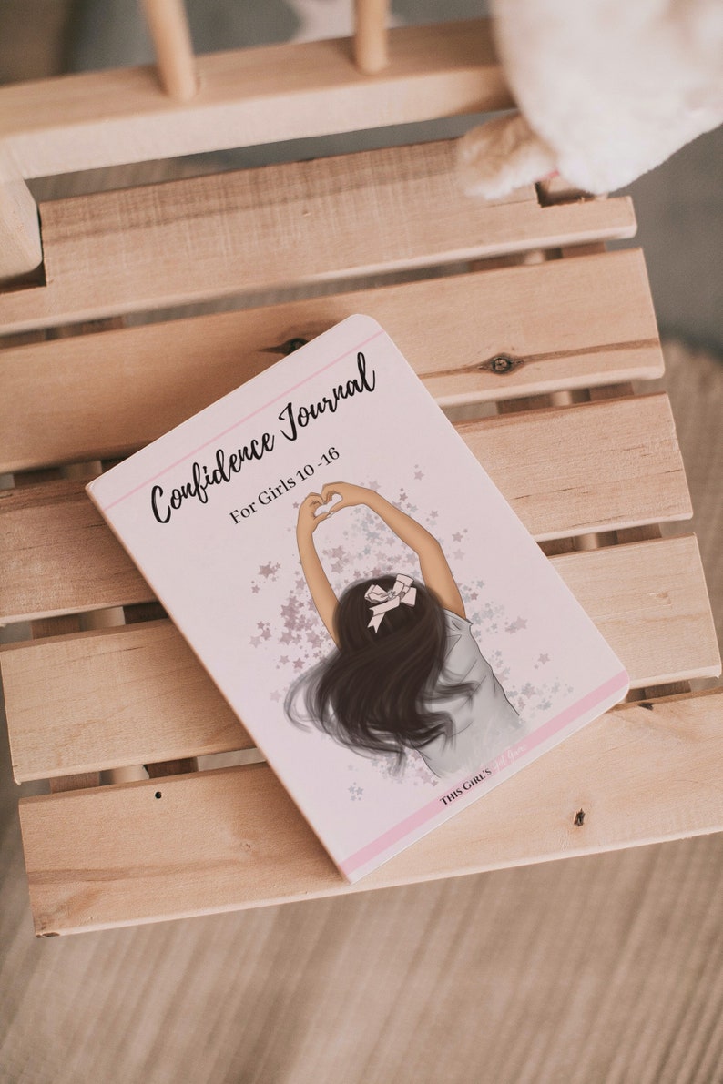 May include: Pink and white journal with the title "Confidence Journal for Girls 10-16" and an illustration of a girl with long brown hair making a heart shape with her hands. The background is a light pink with white stars.
