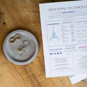 Puede incluir: Una mesa de madera con una bandeja de hormig&oacute;n gris con pendientes dorados y una hoja de trucos de t&eacute;cnicas de conexi&oacute;n a tierra. La hoja de trucos incluye texto y diagramas para ejercicios de respiraci&oacute;n y relajaci&oacute;n.