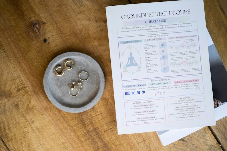 Puede incluir: Una mesa de madera con una bandeja de hormig&oacute;n gris con pendientes dorados y una hoja de trucos de t&eacute;cnicas de conexi&oacute;n a tierra. La hoja de trucos incluye texto y diagramas para ejercicios de respiraci&oacute;n y relajaci&oacute;n.
