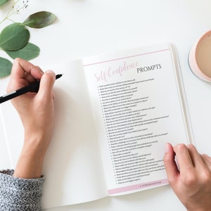 Puede incluir: Cuaderno abierto con una cubierta rosa y las palabras "Self-Confidence Prompts" escritas en &eacute;l. El cuaderno est&aacute; lleno de una lista de preguntas para ayudar a construir la confianza en uno mismo.