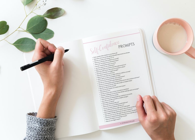 Puede incluir: Cuaderno abierto con una cubierta rosa y las palabras "Self-Confidence Prompts" escritas en &eacute;l. El cuaderno est&aacute; lleno de una lista de preguntas para ayudar a construir la confianza en uno mismo.