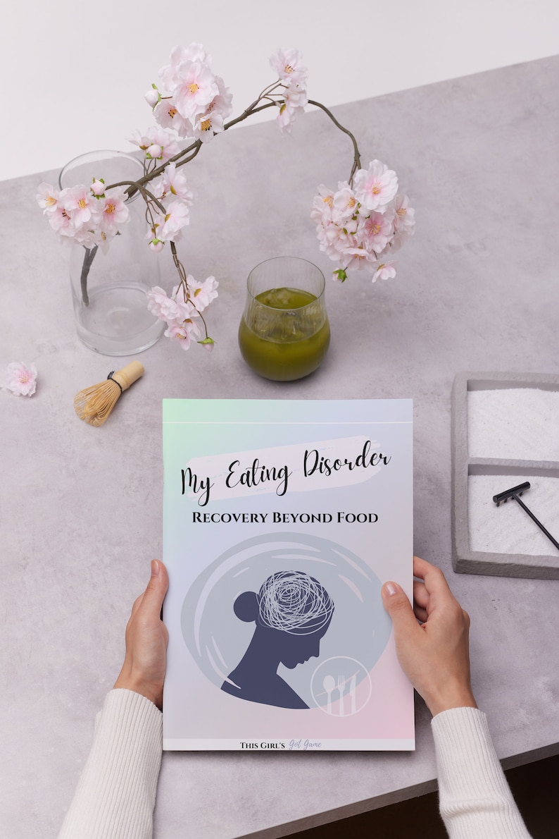 May include: A book titled "My Eating Disorder: Recovery Beyond Food" is held in front of a light grey background. The cover features a silhouette of a person's head with a tangled design, a fork, and a knife. A glass of green liquid and pink flowers are also visible.