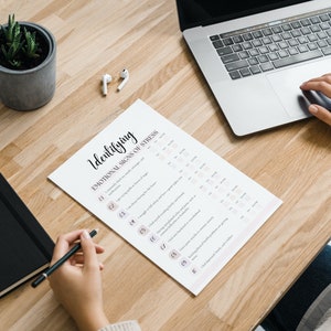 Peut inclure: Une personne est assise &agrave; un bureau avec un ordinateur portable, un carnet, une plante en pot et une feuille de papier avec le titre "Identifier les signes &eacute;motionnels du stress".