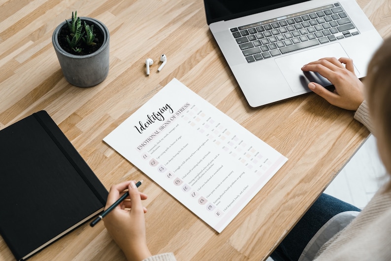 Peut inclure: Une personne est assise &agrave; un bureau avec un ordinateur portable, un carnet, une plante en pot et une feuille de papier avec le titre "Identifier les signes &eacute;motionnels du stress".